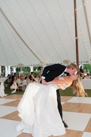 Groom dips bride for a romantic kiss on checkered dance floor under a white tent with string lights.