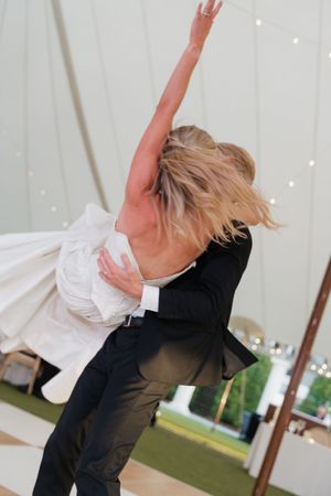 Groom lifting bride in elegant first dance under tent at outdoor wedding reception.