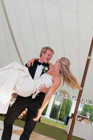 Groom lifts and spins bride in white wedding gown during joyful first dance under a white tent.