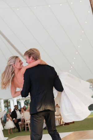 Groom lifting bride during first dance at elegant tented wedding reception with guests seated in background.