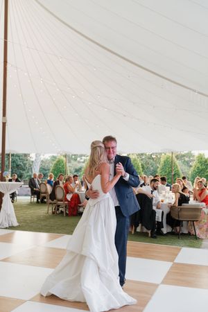 Bride and groom share first dance on checkered floor under elegant white tent with string lights at wedding reception.