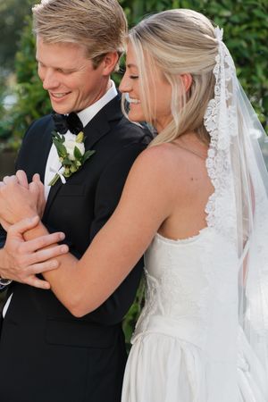 Bride and groom sharing a joyful dance outdoors, bride in lace wedding dress with veil, groom in black tuxedo with boutonniere.