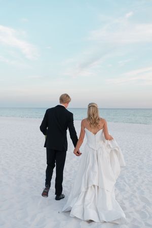 Bride and groom holding hands walking toward the ocean on a white sand beach at sunset.