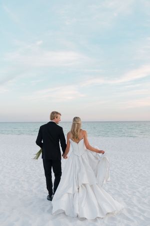Bride and groom walk hand-in-hand along a white sand beach at sunset, bride wearing a layered white gown.