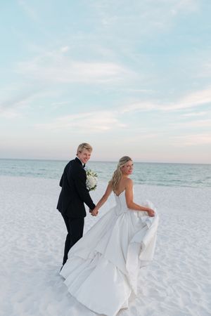 Bride and groom walk hand in hand on a white sand beach at sunset after wedding.