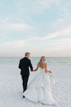 Bride and groom walking hand-in-hand on a white sand beach at sunset in formal wedding attire.