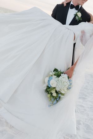Bride and groom embrace on the beach with a blue and white hydrangea wedding bouquet.