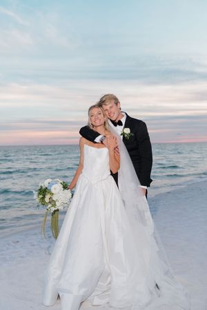 Bride and groom embrace on a beach at sunset, she holds white bouquet in white gown, he wears black tuxedo.