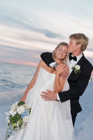 Bride and groom embrace on a beach at sunset, groom kissing bride as she holds a white and blue bouquet.