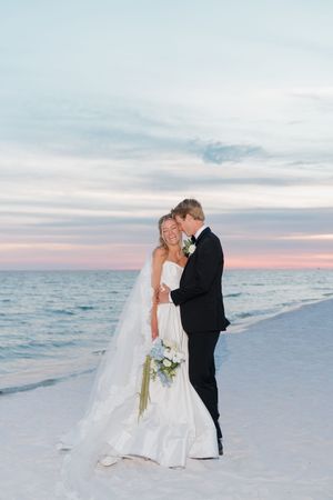 Bride and groom embrace on sandy beach at sunset, bride holding white bouquet with flowing veil trailing behind.