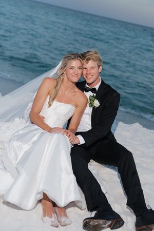 Bride and groom sitting together on white sand beach, smiling, bride in strapless gown with veil, groom in black tuxedo.