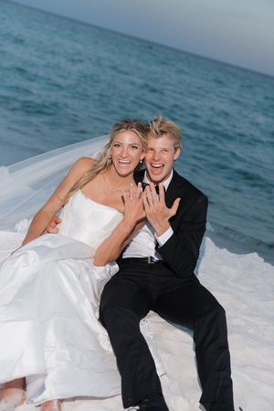 Joyful newlyweds sitting on white sand by the ocean showing off their wedding rings.