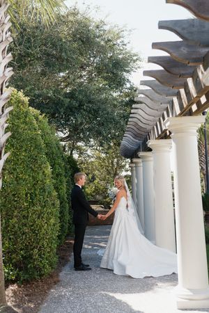 Bride and groom holding hands under a white pergola surrounded by lush hedges and tall trees at their wedding venue.