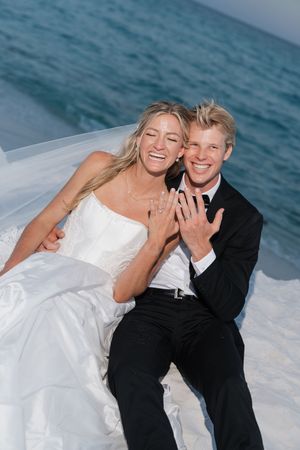 Laughing newlyweds sit on white sand by the ocean showing off their wedding rings in a joyful beach photo.