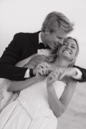 Black and white photo of a groom embracing his laughing bride from behind, holding hands at their wedding.
