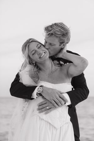 Black and white photo of groom kissing smiling bride on cheek at beach wedding golden hour portrait.