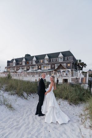 Bride and groom hold hands on white sand beach at dusk with elegant coastal wedding venue in background.