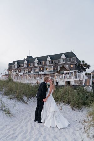 Bride and groom kiss on white sand beach dunes in front of charming coastal hotel at dusk.