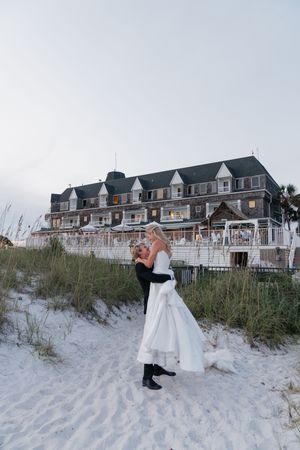 Romantic bride and groom embracing on sandy beach at dusk with a large coastal shingle-style hotel in the background.