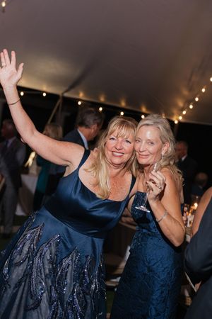 Two women in navy blue dresses celebrate joyfully under string lights at elegant tented wedding reception.