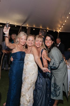 Four women pose together at a wedding reception under a tent with string lights, one holding a champagne glass.