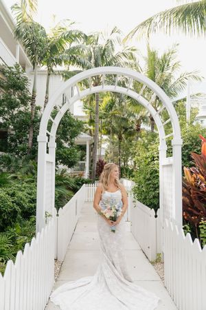 A bride walks under white garden arches surrounded by tropical palm trees and lush landscaping.