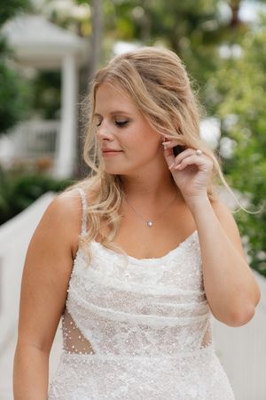 A bride in an elegant white lace wedding dress with blonde hair touches her ear while looking down in an outdoor setting.