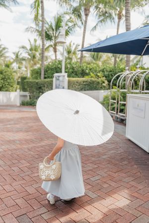 A person holding a white parasol walks on brick pavement near tropical palm trees.