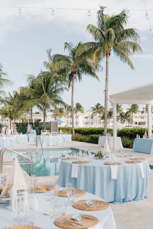 Elegant outdoor wedding reception setup with light blue linens and palm trees by a pool.
