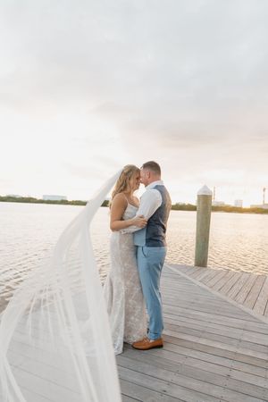 A couple embraces on a wooden dock at sunset with a flowing veil in the breeze.