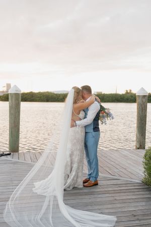 A romantic wedding couple embraces on a wooden dock at sunset with water in the background.
