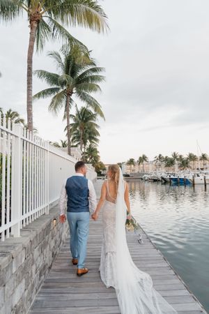 Couple walking along a waterfront boardwalk with palm trees and white railings.