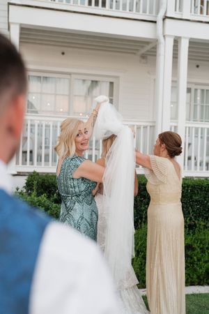 Wedding party members help adjust a long white veil on a porch.