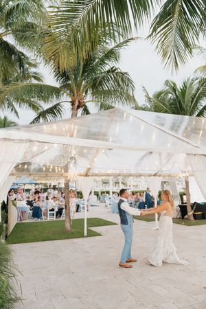 Wedding reception setup under clear tent with palm trees and string lights at dusk.