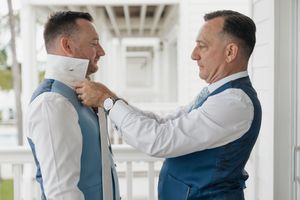Groomsmen in navy suits and white shirts help each other prepare for a wedding ceremony.