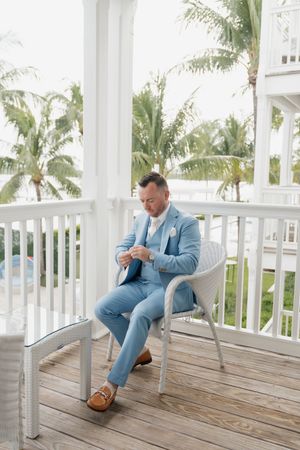 A light blue suited figure seated on a white chair on a tropical veranda with palm trees.