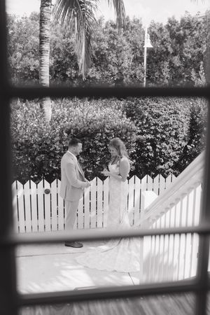 A black and white photo of a couple in wedding attire standing by a white picket fence.