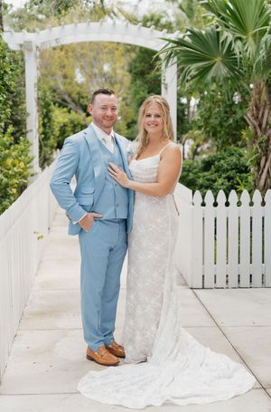 A couple poses under a white garden arbor with palm trees and picket fence in the background.
