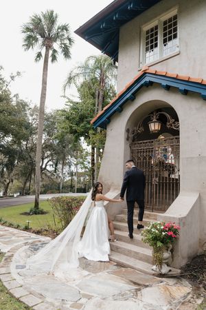 A couple in formal attire enters through an ornate archway at a Spanish-style venue with palm trees and stone pathway.