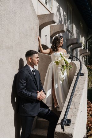 A wedding couple poses on stone steps with dramatic shadows as the bride holds a white floral bouquet.