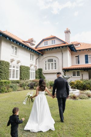 A couple in wedding attire walks hand in hand towards a grand white mansion with a small child following behind.