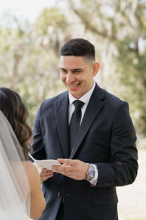 A groom in a black suit reads his vows from a paper while smiling during an outdoor wedding ceremony.