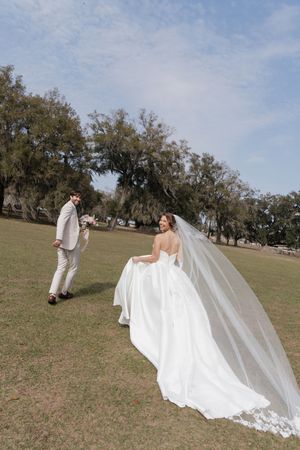 A wedding couple walks across a grassy field with flowing veil and oak trees in the background.
