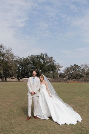 A wedding couple poses in an open field with trees, the bride's white dress flowing in the breeze.