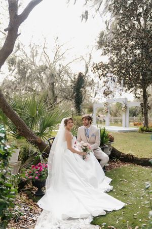 A wedding couple sits on a garden bench with palm trees and a white gazebo in the background.