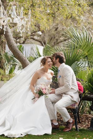 A romantic wedding couple shares tender moments while sitting on a garden bench surrounded by lush greenery.