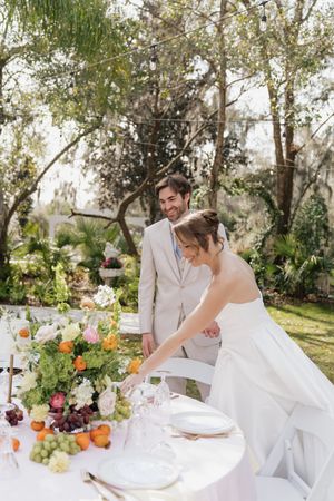 A couple stands at their outdoor wedding reception table decorated with fresh flowers and fruit.