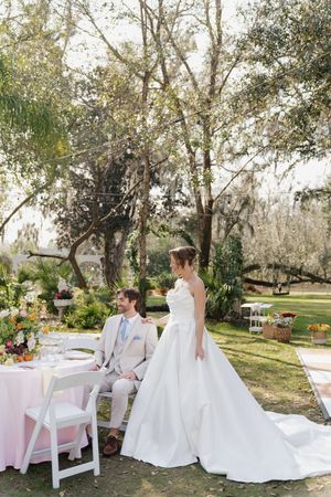 An outdoor wedding reception setup with white furniture and decor under a canopy of trees.