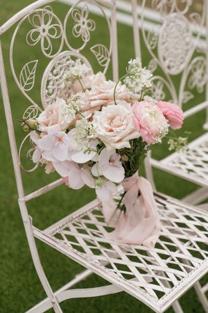 A delicate bouquet of pink roses and baby's breath sits on a white ornate metal garden chair against a green background.