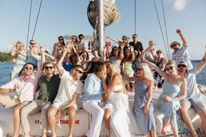 Large group of friends celebrating on a sailboat, raising glasses and cheering together on a sunny day.